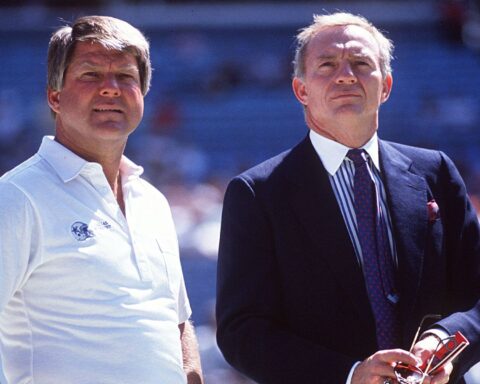 Two men standing outdoors, one in a white shirt and the other in a suit, with a blurred crowd in the background.