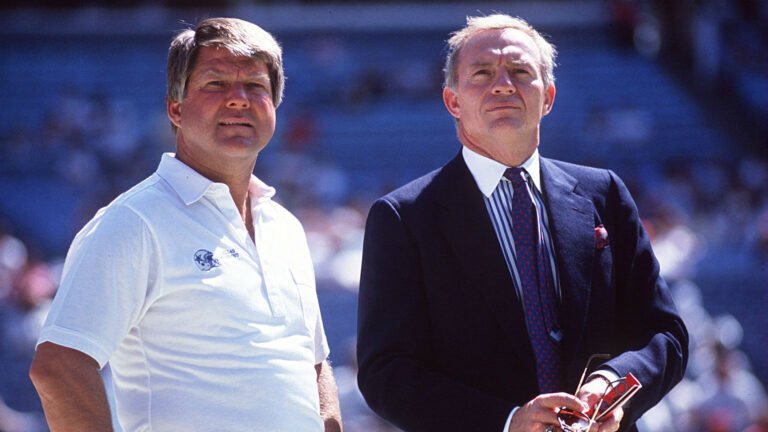 Two men standing outdoors, one in a white shirt and the other in a suit, with a blurred crowd in the background.