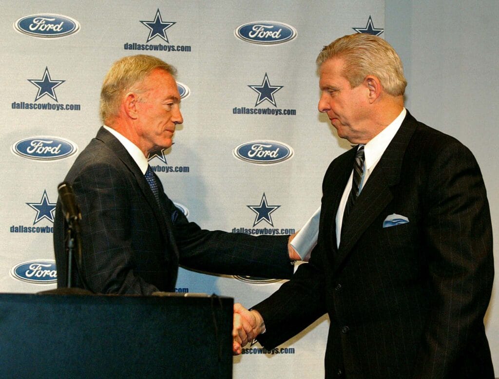 Jerry Jones has specific reasons for choosing Brian Schottenheimer 1 Two men in suits shake hands in front of a Dallas Cowboys backdrop with Ford logos.