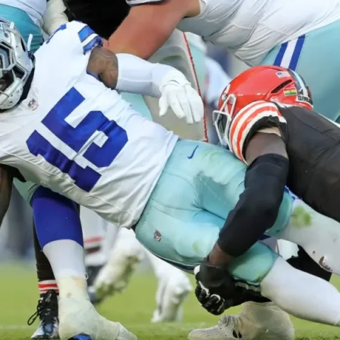 During the game, a Cowboys player in a blue and white uniform is tackled by an opponent in black and orange, showcasing intense action despite his decreasing role before the bye week.