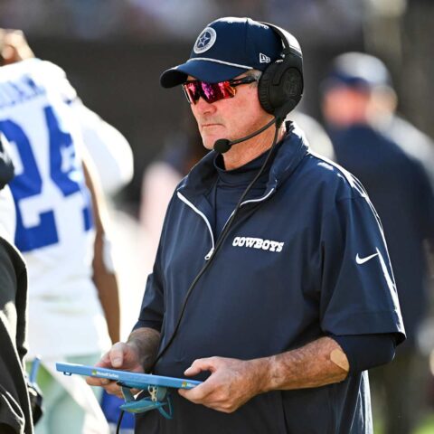A football coach for the Cowboys, sporting sunglasses and a headset, looks at a tablet on the sidelines during a game this season.