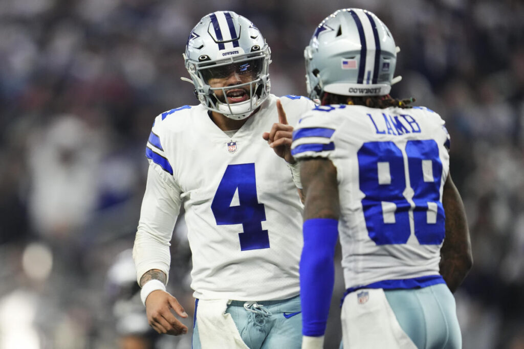 Two football players in white Dallas Cowboys uniforms, adorned with stars, communicate on the field during a game.