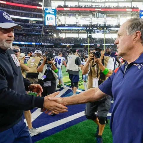 Two football coaches shake hands on the field after a game, surrounded by photographers and staff.