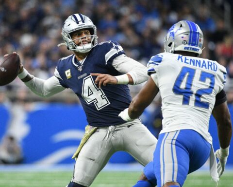 Football player in white and blue uniform throws a pass as a defender in white and blue approaches.