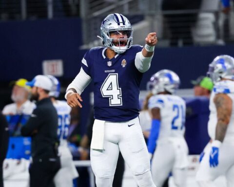 Football player in a blue uniform points forward during a game, surrounded by team members on the field.