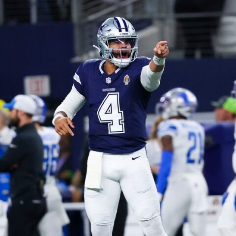 Football player in a blue uniform points forward during a game, surrounded by team members on the field.