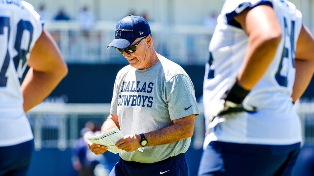 A coach in sunglasses and a Dallas Cowboys shirt reads a paper during a practice, with players nearby.