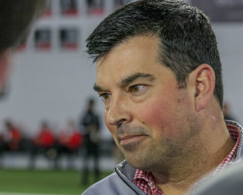 A man in a gray jacket and red checkered shirt looks to the side indoors, exuding the steady focus of a coaching staff member.