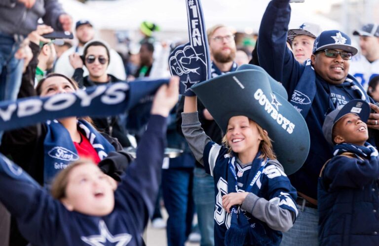 Cowboys fans in team gear cheer enthusiastically at a sports event, waving foam fingers and banners.