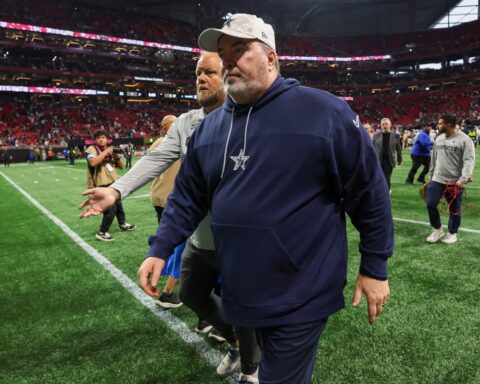 A man in a navy tracksuit and cap walks on a football field, reminiscent of a Dallas Cowboys practice session, with people and stadium seats in the background.