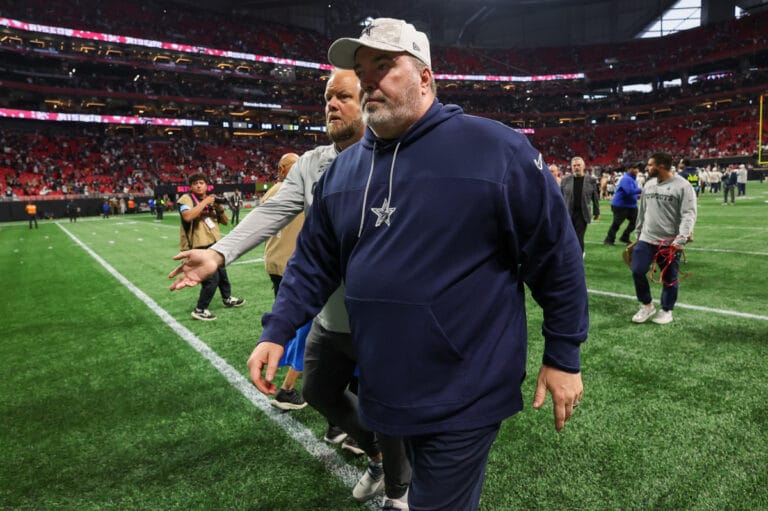 A man in a navy tracksuit and cap walks on a football field, reminiscent of a Dallas Cowboys practice session, with people and stadium seats in the background.