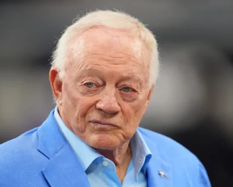 A man in a light blue blazer, reminiscent of Jerry Jones' favored style, gazes ahead with a neutral expression, embodying the stoic demeanor of a seasoned Cowboys head coach.