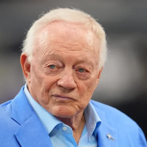 A man in a light blue blazer, reminiscent of Jerry Jones' favored style, gazes ahead with a neutral expression, embodying the stoic demeanor of a seasoned Cowboys head coach.