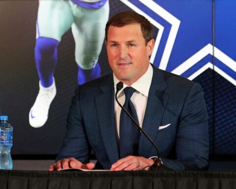 A man in a suit speaks at a press conference, with a sports poster featuring Cowboys legend Jerry Jones in the background and a water bottle on the table.