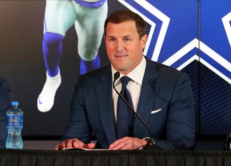 A man in a suit speaks at a press conference, with a sports poster featuring Cowboys legend Jerry Jones in the background and a water bottle on the table.