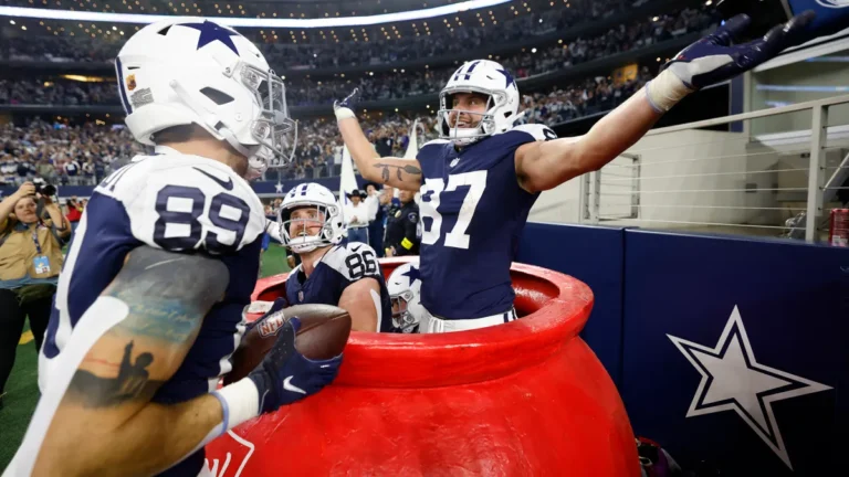 During a Thanksgiving game in the 2024 season, a Cowboys football player celebrates exuberantly in a large red bucket on the sidelines, with two teammates laughing nearby.