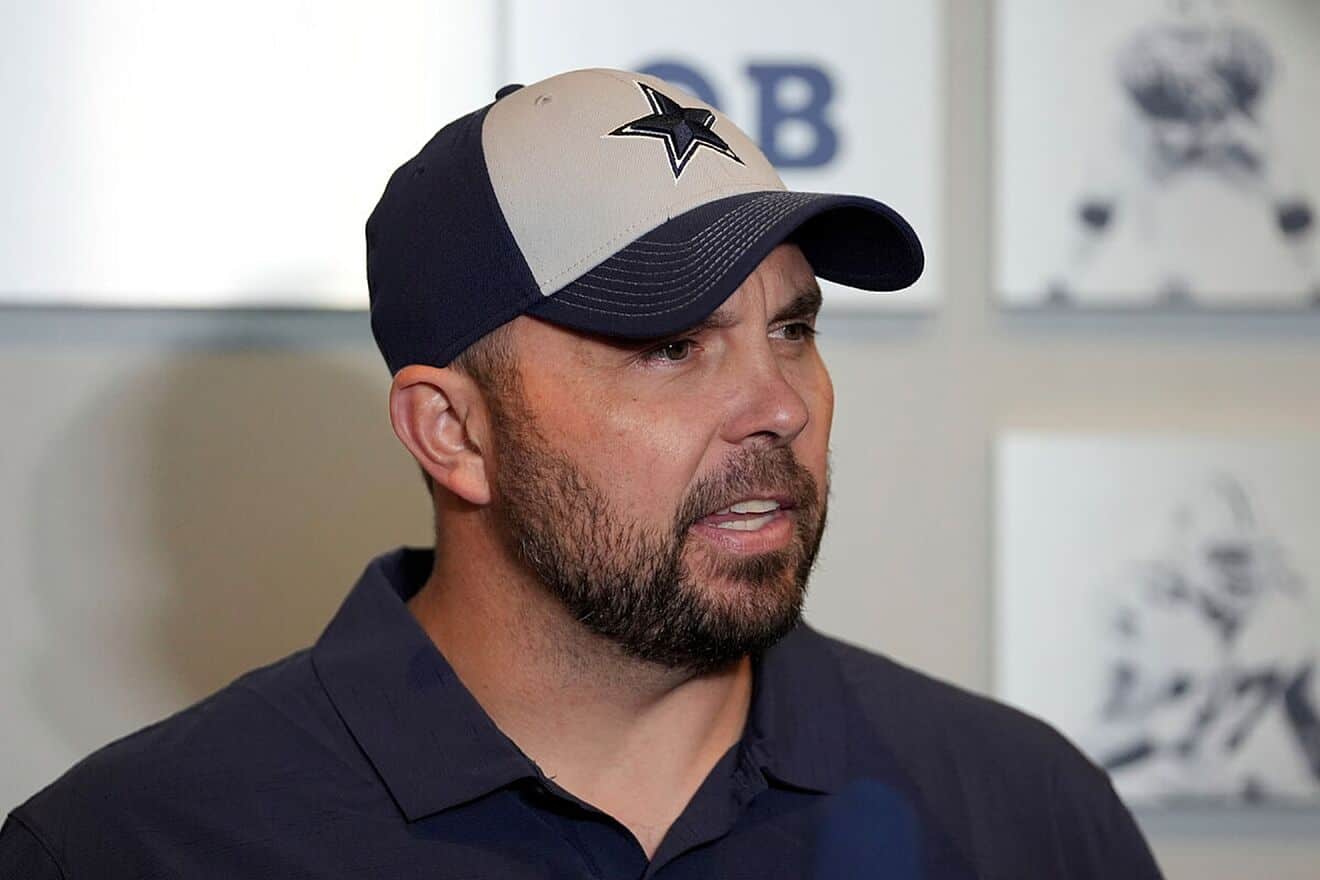 Dallas Cowboys offensive coordinator Klayton Adams speaking to the media. He is wearing a white and blue baseball cap with a blue star in the center of the hat. Breece Hall to the Cowboys: Trade Scenario, Stats, and 2025 Impact
