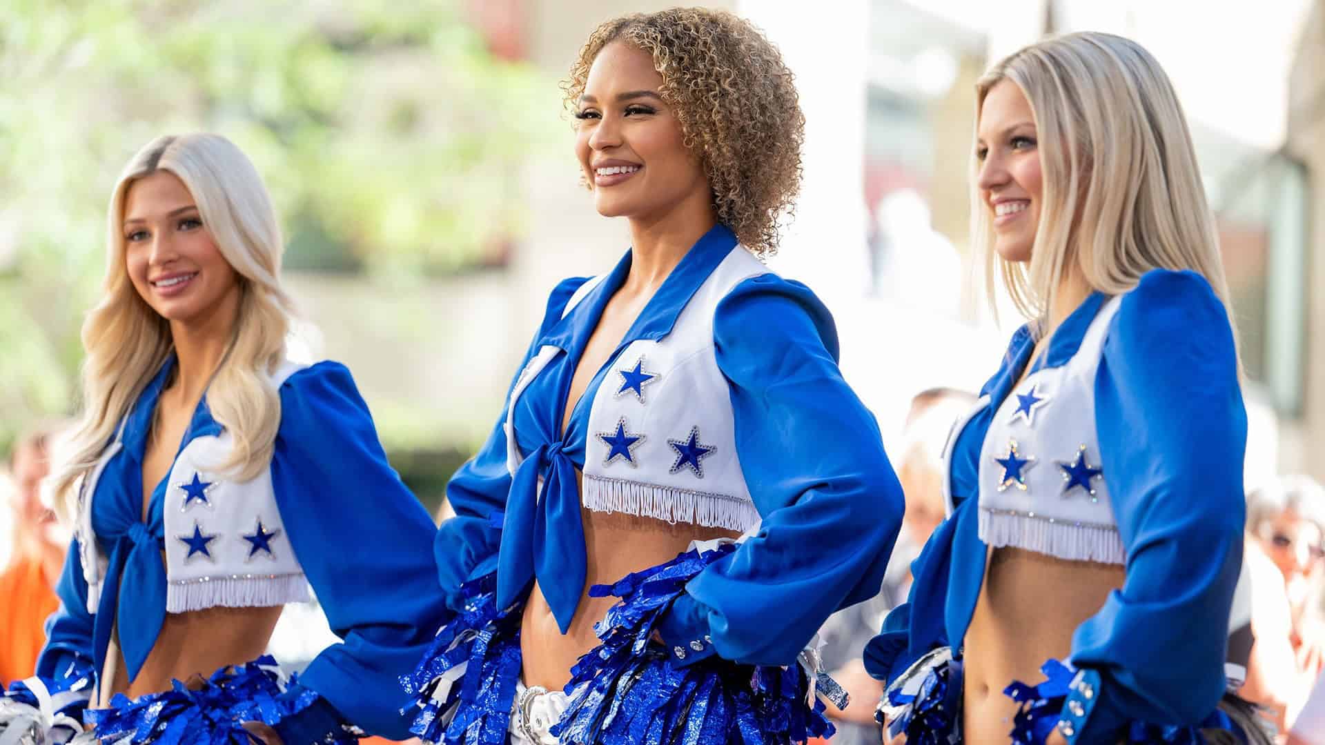 Three Dallas Cowboys Cheerleaders in white vest with blue stars adorn to the front and a blur long sleeve shirt underneath the vest. Cowboys Cheerleaders: Netflix Sparked a 400% Raise