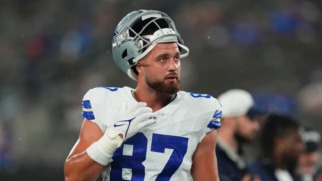 Dallas Cowboys Tight End Jake Ferguson in a white Dallas Cowboys jersey with his helmet resting on the top of his head. Tight End Battle: 2025 Cowboys Could See a Shake-Up