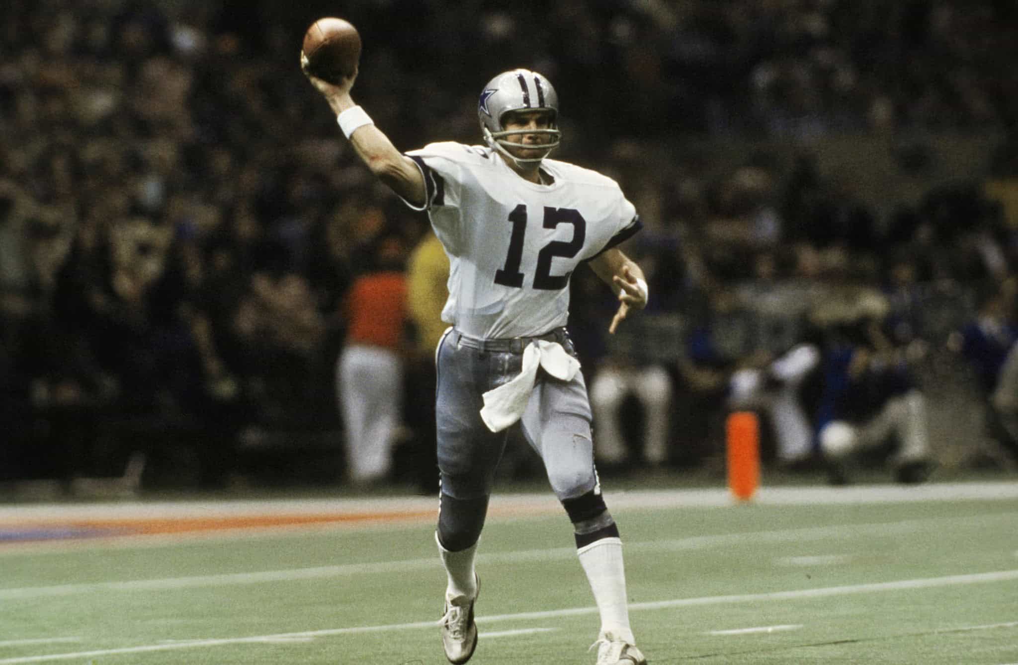 Roger Staubach, wearing a white number 12 Dallas Cowboys jersey, throwing a football. The GOAT of the Dallas Cowboys