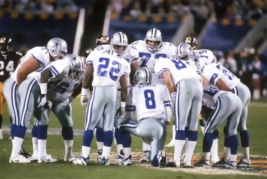 The 1992 Dallas Cowboys offense huddled around Troy Aikman who is giving the play call. How No Salary Cap Fueled the Cowboys' 90s Dynasty